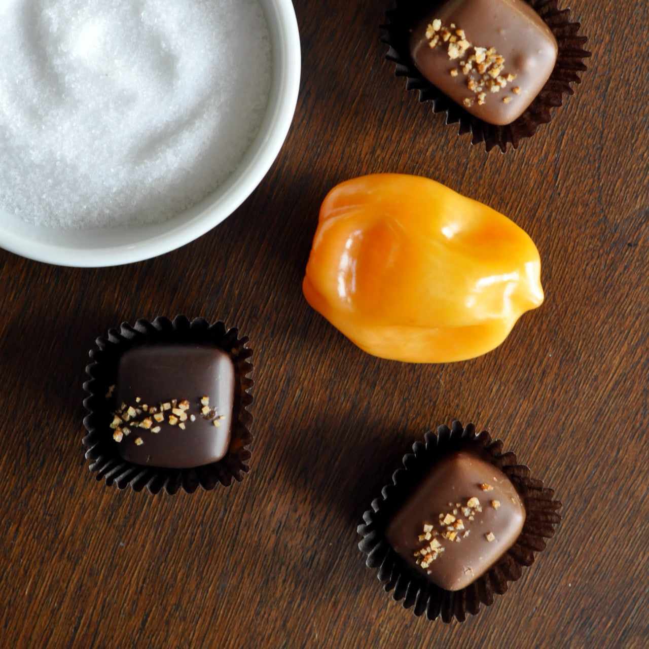 Three chocolate candies with sea salt and habanero sprinkled on top with a fresh habanero pepper and bowl of sea salt next to them